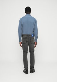 Light blue long-sleeve shirt with buttoned cuffs and dark gray slim-fit jeans. Black shoes complete the outfit. Rear view with brand label visible.