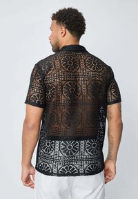 Chemise noire en crochet à manches courtes avec un motif floral et une coupe décontractée, présentant une texture en maille ouverte et un col à boutons.
