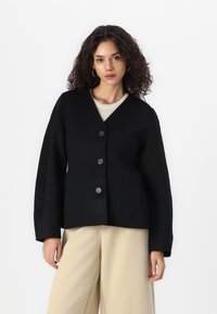Svart ullblanding jakke med V-hals, fire store knapper og strukturerte ermer. Båret over en kremfarget strikket topp og beige bukser.