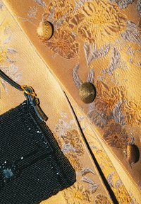 Tessuto dorato con motivo floreale, superficie testurizzata e bottoni prominenti, accanto a una piccola borsa a tracolla nera con perline.