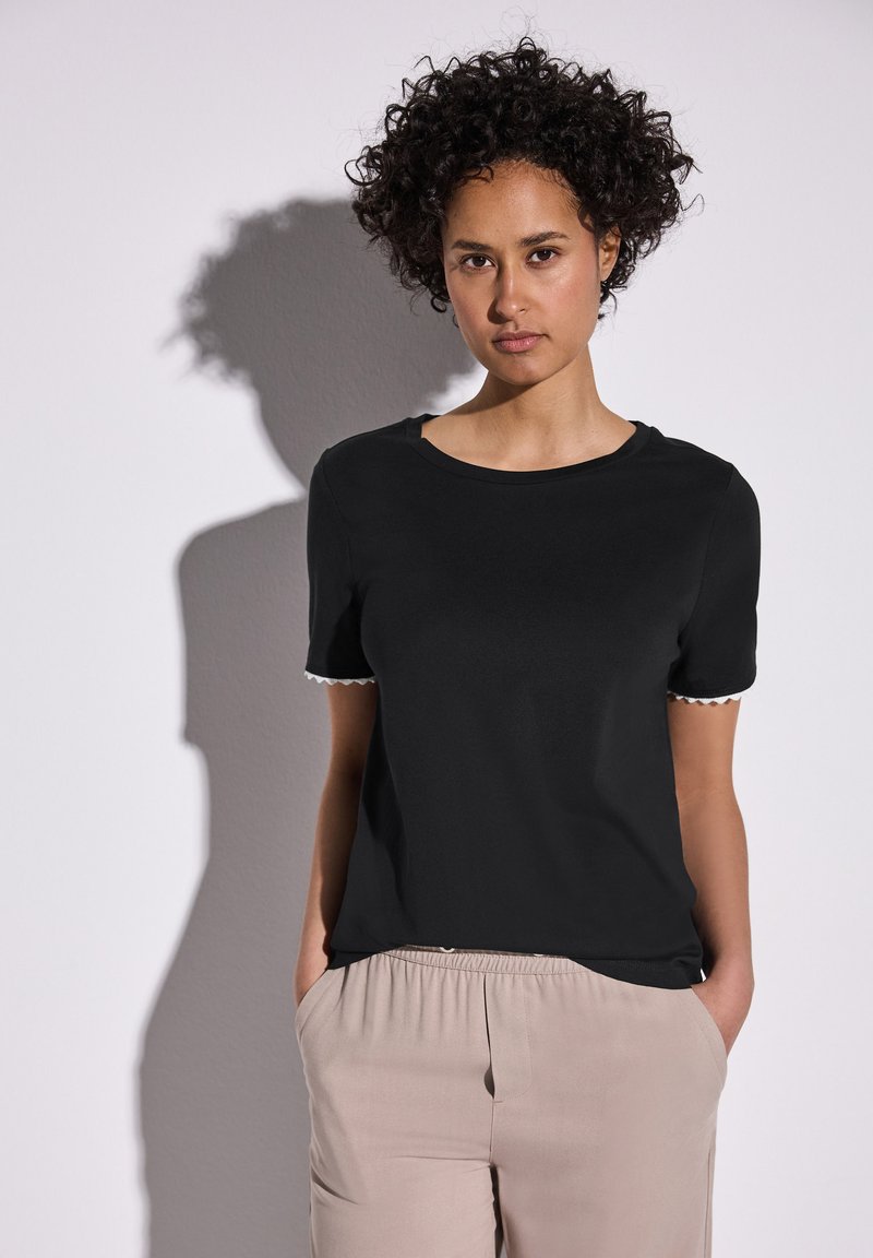 Mustat lyhythihaiset t-paidat, joissa on valkoiset aaltoilevat yksityiskohdat hihoissa, valmistettu sileästä kankaasta. Yhdistetty vaaleanbeigeihin, löysiin housuihin.