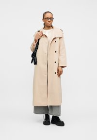 Γυναίκα με πλεγμένα μαλλιά, φοράει γυαλιά, μπεζ μακρύ παλτό, γκρι τζιν με φαρδιά μπατζάκια, μαύρες μπότες, κρατώντας μαύρη τσάντα ώμου σε λευκό φόντο.