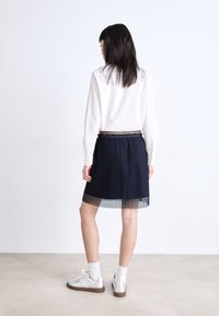 Camisa blanca de manga larga con rayas verticales, falda plisada marina con un fondo transparente, acompañada de zapatillas plateadas y calcetines blancos.