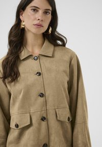 Jachetă din suede maro, cu guler, cinci butoane și două buzunare frontale. Textură moale, cu un design confortabil.