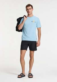 T-shirt di cotone azzurro con stampa, abbinata a pantaloncini neri e sandali grigi. L'uomo tiene una giacca nera sulla spalla.