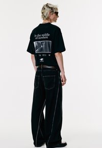 Persona con capelli biondi corti e occhiali da sole indossa una maglietta nera con grafica con il testo "In the middle of nowhere" e pantaloni neri larghi, in piedi di profilo.