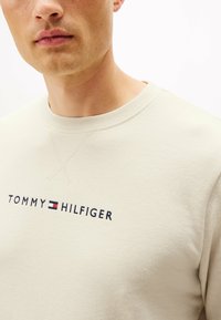 Jasno beżowy sweatshirt z prążkowanym okrągłym dekoltem, widocznymi detalami szwów i logiem "TOMMY HILFIGER" haftowanym ciemnonawym.