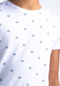 Biała koszulka z powtarzającymi się wzorami w kolorze teal przedstawiającymi słońce i fale. Materiał wydaje się być miękki i lekki, z okrągłym dekoltem i krótkimi rękawami.