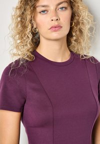 Kurzärmliges lila Oberteil aus glattem Stoff mit Rundhalsausschnitt und dezenten Nahtdetails, das eine figurnahe Silhouette betont.