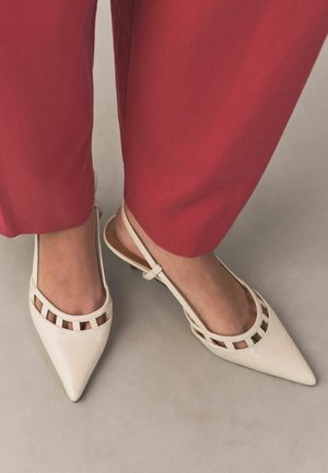 Jalad, mis kannavad valgeid terava ninaga rihmikkingi lõhikdetailidega, ühendatuna punaste laia säärega pükstega heleda halli põranda peal.