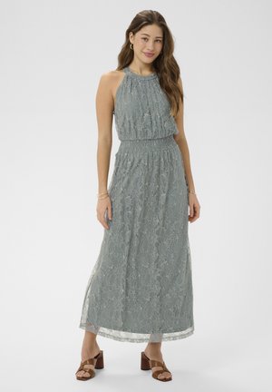 Žena v sivých čipkovaných maxi šatách bez rukávov so stiahnutým pásom a hnedými sandálmi na podpätku stojaca pred jednofarebným pozadím.