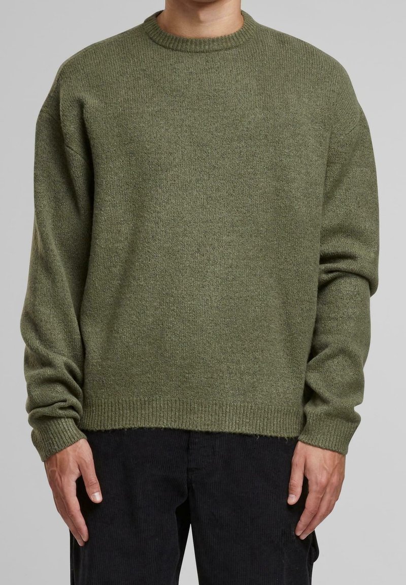 Personne portant un pull en tricot vert olive à col rond avec manches longues et un pantalon noir, debout devant un fond uni.