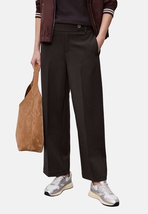 Osoba nosząca ciemne, szerokie spodnie, białe i srebrne trampki oraz bordową kurtkę, trzymająca obok siebie beżową zamszową torbę na ramię.