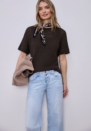 Kobieta ubrana w ciemnobrązowy krótki sweter z krótkim rękawem, jasnoniebieskie dżinsy z wysokim stanem, szalik na szyję w zwierzęcy wzór, trzyma złożoną beżową kurtkę opartą o białą ścianę.