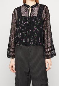 Femme portant un chemisier noir transparent à fleurs avec bordure en dentelle et liens à pampilles, associé à un pantalon cargo noir à fines rayures.