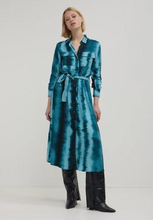 Una donna che indossa un abito camicia in satin turchese e nero con una cintura in vita e stivali di pelle neri al ginocchio, in piedi su uno sfondo neutro.
