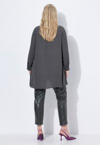 Woman with long blonde hair wearing a loose dark gray tunic, black leather pants, and purple high-heeled slingback shoes, standing against a white wall.