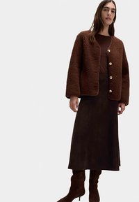 Veste marron texturée avec de grands boutons dorés, associée à une longue jupe marron et des bottes marron montant jusqu'aux genoux.