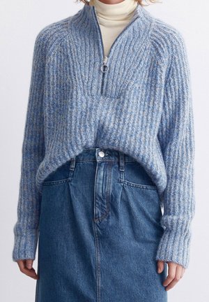 Pull en maille côtelée bleu et gris avec une demi-fermeture éclair, porté sur un col roulé crème, associé à une jupe en jean bleu taille haute.