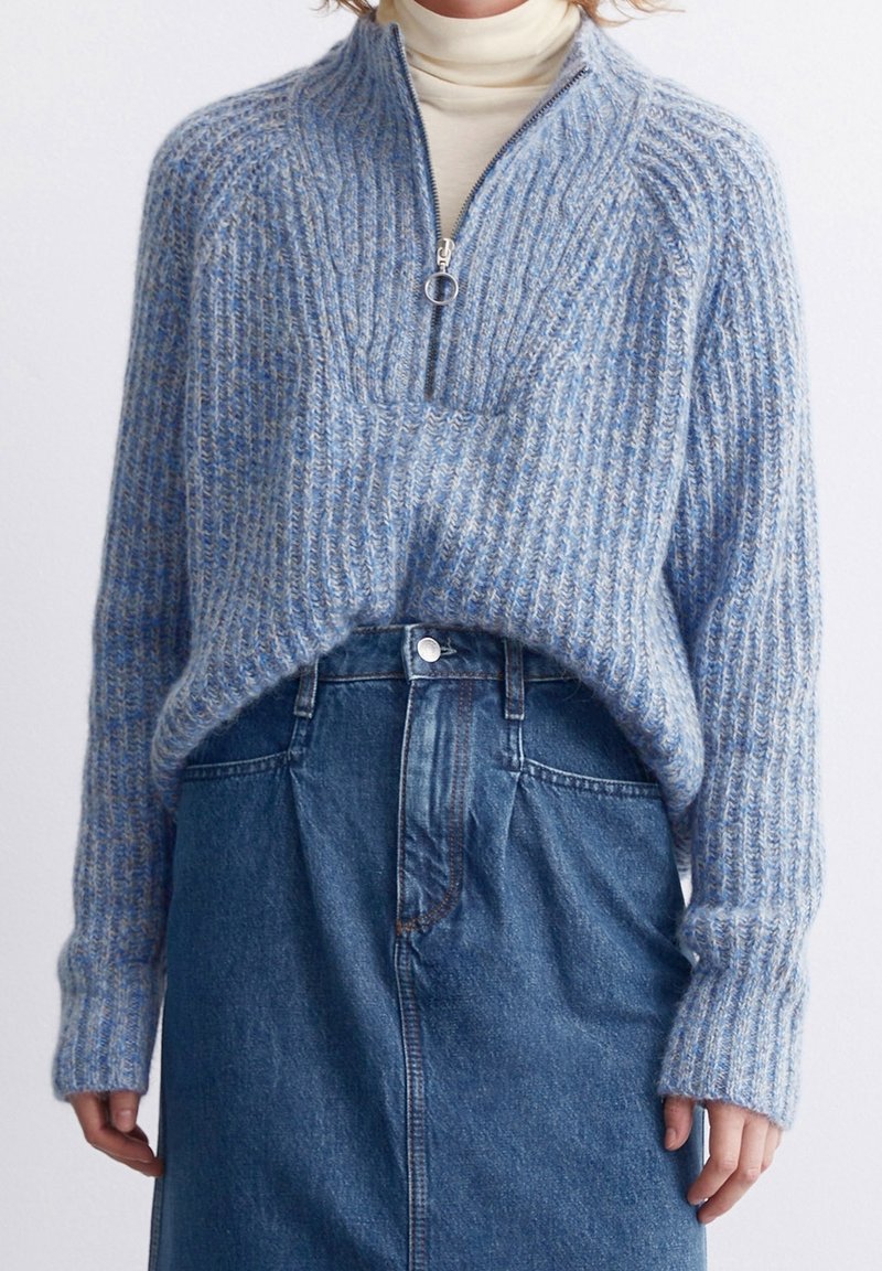 Pull en maille côtelée bleu et gris avec une demi-fermeture éclair, porté sur un col roulé crème, associé à une jupe en jean bleu taille haute.