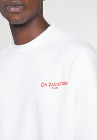 Bílá mikina s žebrovaným kulatým výstřihem, s nápisem "ON VACATION CLUB" v korálově růžové barvě na hrudi. Hladká textura tkaniny.