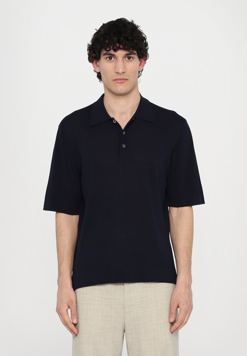 Polo à manches courtes bleu marine avec un col plat et une patte de boutonnage à trois boutons, en tissu lisse. Associé à un pantalon léger et texturé.