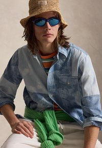 Tânăr care poartă ochelari de soare cu lentile albăstrui, o pălărie din paie tip bucket, un tricou albastru cu imprimeu tie-dye, un tricou cu dungi și un pulover verde legat în jurul taliei.