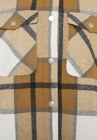 Camicia a quadri con colori marrone, crema e nero, realizzata in tessuto morbido e texture. Include bottoni metallici e due tasche sul petto.