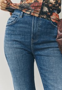 Tmavomodré džínsy z denimu s klasickým dizajnom s piatimi vreckami. Viditeľné švy a hladká textúra. Na páse je čiastočne prekrytá vzorovaná kvetinová blúzka.