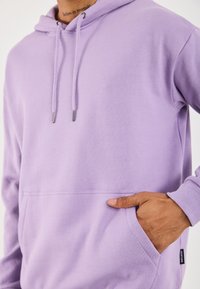 Hoodie viola chiaro con coulisse regolabile, tasca frontale, polsini a coste e texture in felpa morbida. Presenta un'etichetta con logo sull'orlo.