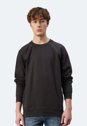 Giovane uomo con capelli ricci che indossa una felpa nera a maniche lunghe semplice e jeans blu, in piedi contro uno sfondo grigio chiaro.