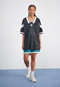 Black mesh oversized jersey with white accents and logo. Teal shorts with white lace trim. Model wears white socks and light sneakers.