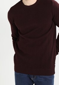 Burgundy texturerad tröja med fiskbensmönster, rund hals, och ribbade ärmslut och fåll. I kombination med blå jeans.