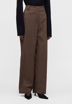 Pantalon marron à jambes larges porté avec un haut noir à manches longues et des chaussures noires à talons aiguilles sur un fond uni.
