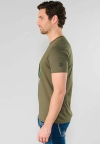 Camiseta de algodón verde oliva con mangas cortas, cuello redondo y un estampado gráfico en el frente. Presenta un pequeño logo en la manga.