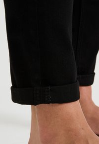 Jambes inférieures portant un pantalon noir retroussé montrant la cheville et les surpiqûres de l'ourlet en détail clair sur un fond uni.