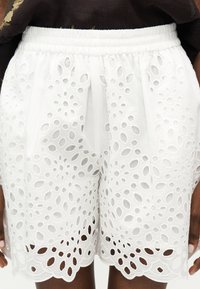 Λευκά σορτς με ελαστική μέση, στολισμένα με κεντήματα από λουλουδένιο ντεκολτέ και κυματιστή άκρη, φορεμένα από ένα άτομο με σκουρόχρωμο δέρμα και σκούρα καφέ μπλούζα.