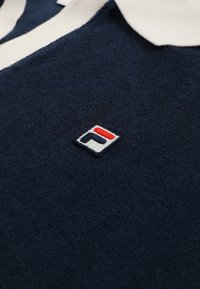 Granatowa, teksturowana tkanina z małym haftowanym logo w kolorach czerwonym, białym i niebieskim oraz widocznym białym obszyciem kołnierzyka.