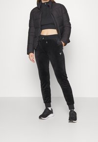 Zwarte pufferjas met een hoge kraag, gecombineerd met zwarte velours joggingbroeken met een elastische tailleband. Zwarte sportschoenen maken de look af.