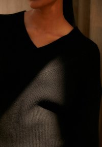 Pull noir en maille avec encolure en V, présentant une texture côtelée et un tissu doux, partiellement illuminé par des motifs d'ombre et de lumière.