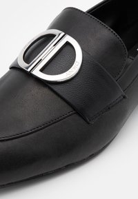 Mocassin en cuir noir avec une large sangle ornée d'une boucle logo en ton argent, texture lisse et design à bout arrondi.