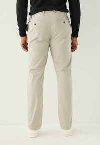 Pantaloni beige chiaro con taglio slim, realizzati in tessuto morbido. Presentano due tasche posteriori con chiusura a bottone. Indossati con una maglietta nera.