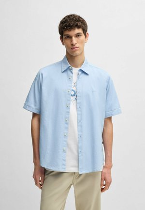 Camisa de manga corta azul claro con cuello abotonado, que presenta una sutil textura y un pequeño logo en el lado izquierdo.
