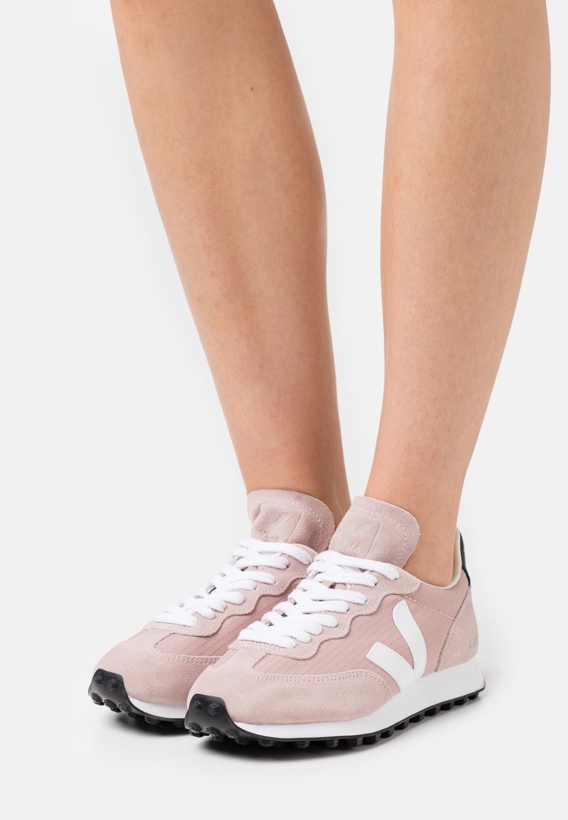Baskets en daim rose avec des lacets blancs et un logo blanc, comportant un accent noir au niveau du talon et une semelle en caoutchouc texturée.