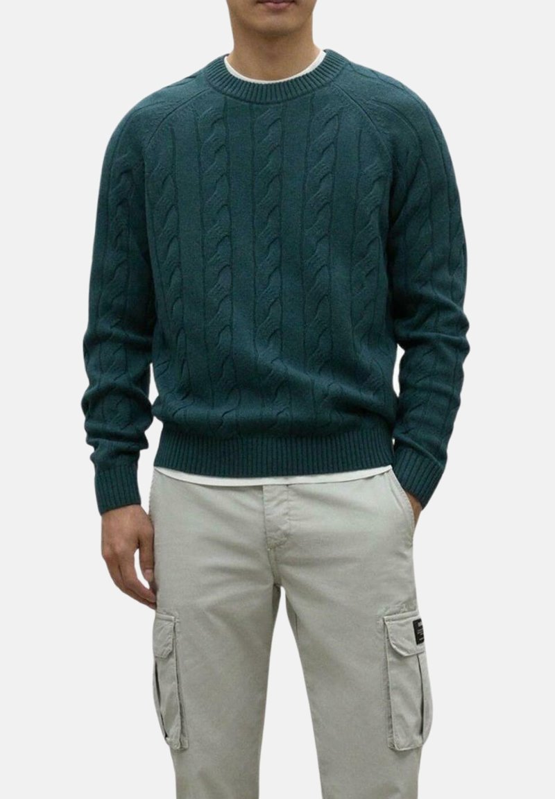 Suéter de punto en color verde azulado con patrón de trenzas, puños y dobladillo acanalados. Lleva puesto sobre una camisa blanca, combinado con pantalones cargo en un tono beis claro.