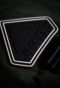 Černá gumová nášivka ve tvaru trojúhelníku s bílým okrajem, s raženým textem a designovými prvky, umístěná na tmavě zeleném textilním pozadí.