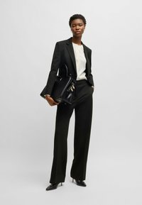 Completo nero su misura con pantaloni larghi, indossato su una blusa crema. Borsa nera con dettagli dorati. Tacchi a punta neri.