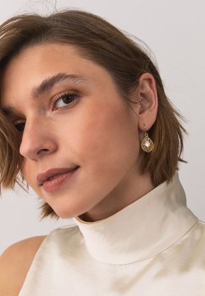 Young woman with short brown hair wearing a white sleeveless turtleneck and a gold teardrop earring with an intricate design.