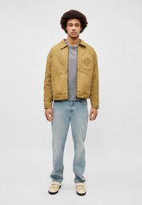 Καφέ βαμβακερό μπουφάν με γιακά και μπροστινή τσέπη, φορεμένο πάνω από γκρίζο πουκάμισο, συνδυασμένο με ανοιχτόχρωμο μπλε τζιν και μπεζ παπούτσια.