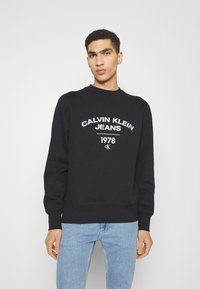 Felpa nera con scollo rotondo e polsini a coste. Presenta testo bianco: "CALVIN KLEIN JEANS 1978" e logo. Indossata con jeans azzurri chiari.
